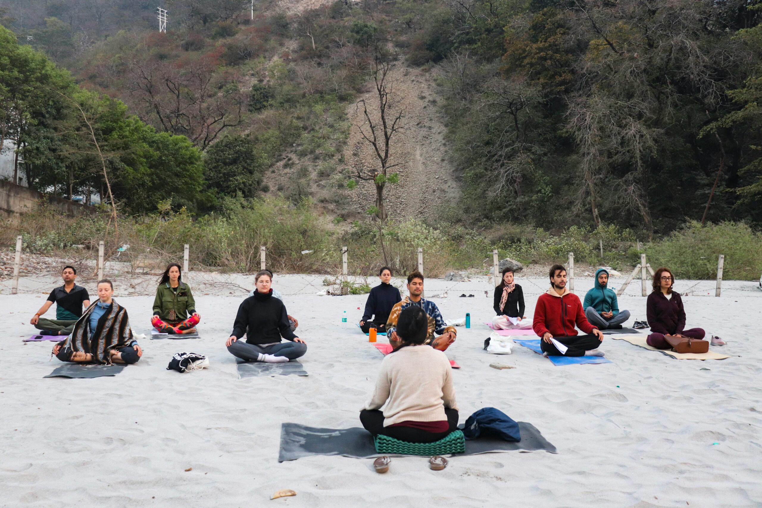 A group practicing meditation on a sandy beach in Rishikesh, India, promoting mindfulness and relaxation.